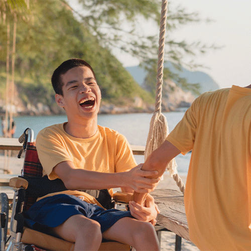 Young person using a wheelchair enjoying time at the beach with support worker, representing inclusive disability and continence care services.